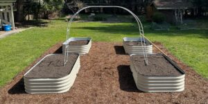 four raised garden beds with white rectangular tin sheets, silver arches in the middle of each bed and walkway between them on top dirt covered grassy backyard with brown wood chips